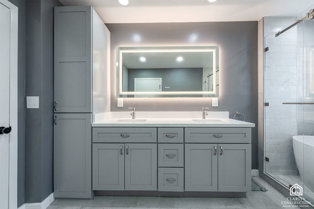 Gray double vanity with a wide backlit mirror and a freestanding tub beyond