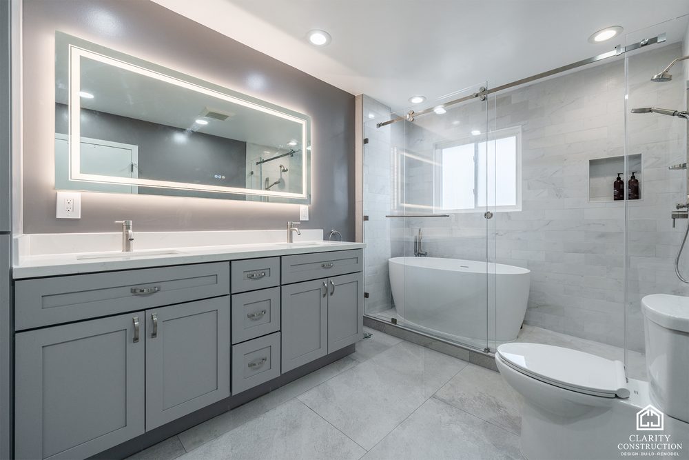 Wide primary bathroom with a gray double vanity, backlit mirror and freestanding tub
