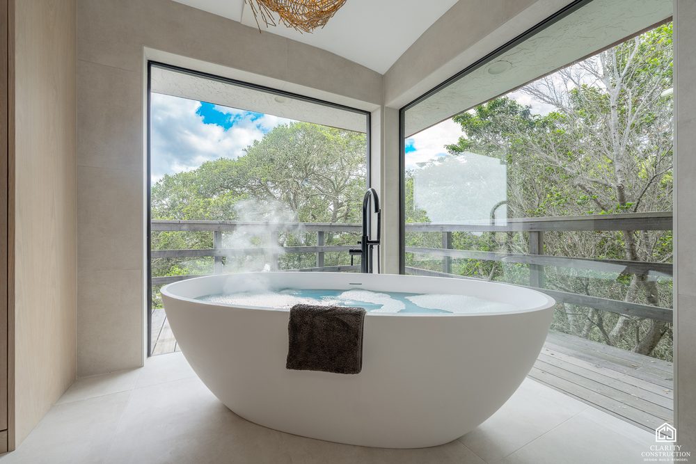 Freestanding soaking tub filled beside a corner window looking into the trees