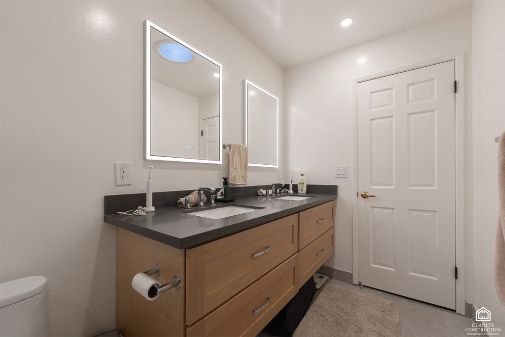 Oak double vanity with a dark stone top and a backlit mirror beside a tub shower