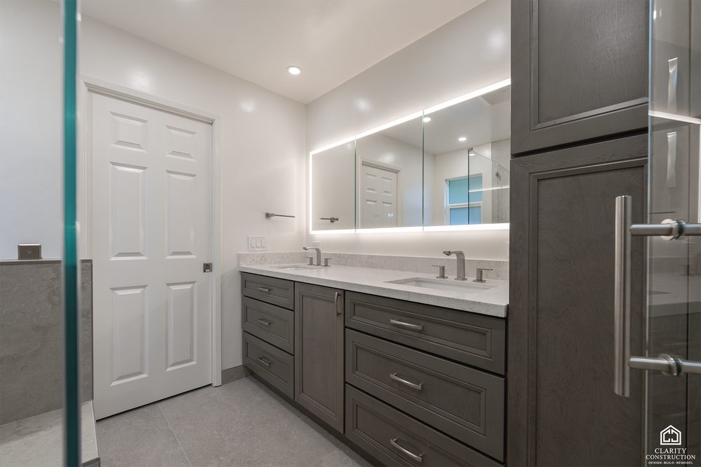 Gray double vanity with a backlit mirror beside a glass fronted shower enclosure