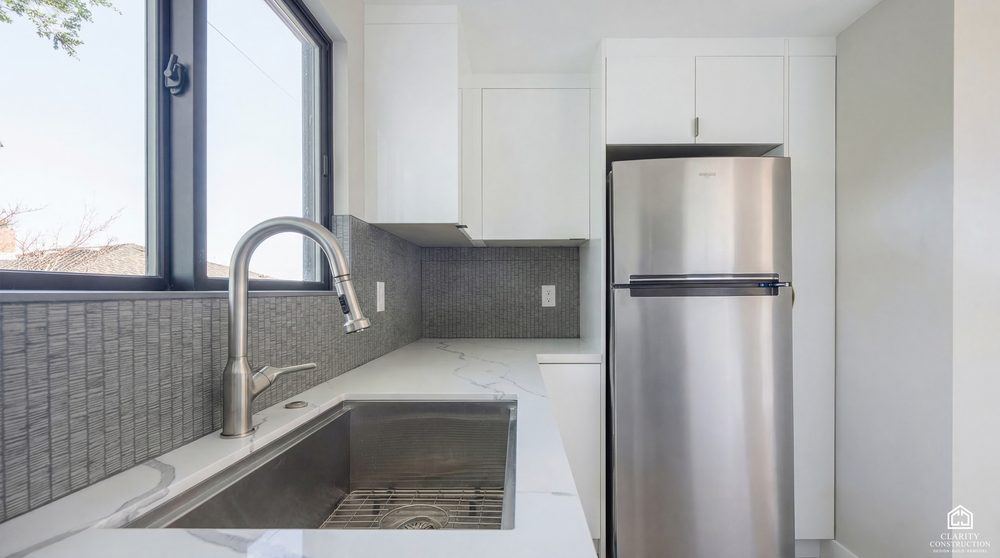 White kitchen corner with a stainless fridge, gray tiled backsplash and a window over the sink