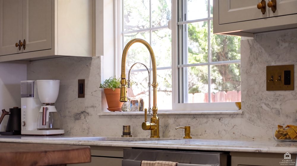 Farmhouse sink under a window with an unlacquered brass faucet and marble slab surround