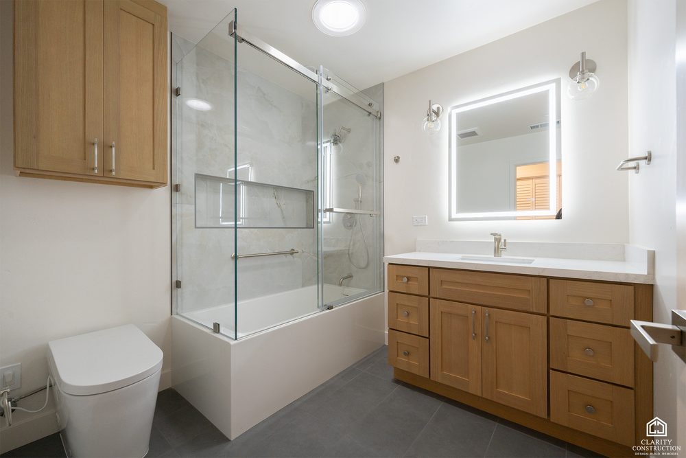 Bathroom with an oak vanity, backlit mirror and a tub shower with a glass screen
