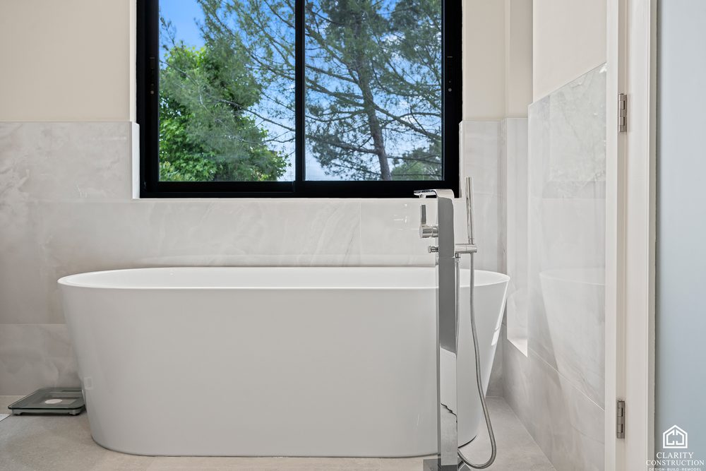 Freestanding tub with a floor mounted filler against a marble wall and a black framed window