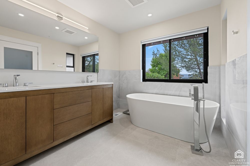 White oak double vanity along a marble wall, with a freestanding tub at the end