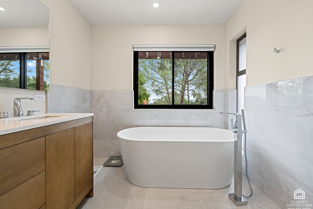 Freestanding tub under a black framed window, with an oak vanity and marble walls