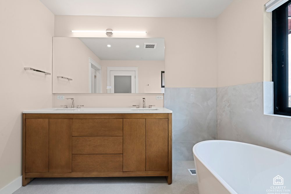 Oak double vanity with a stone top and a freestanding tub beside a marble wall