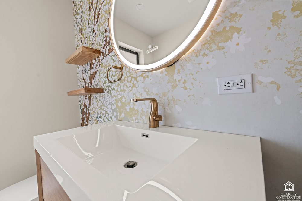 White sink on a floating oak vanity with a brass faucet and wallpaper behind