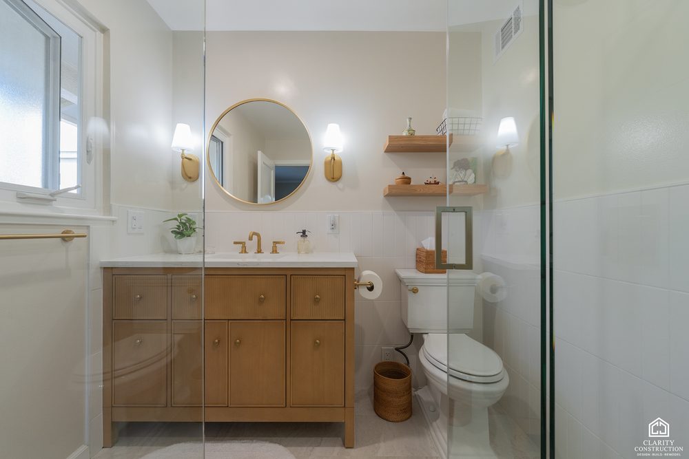 Oak vanity and round mirror seen past the glass shower door