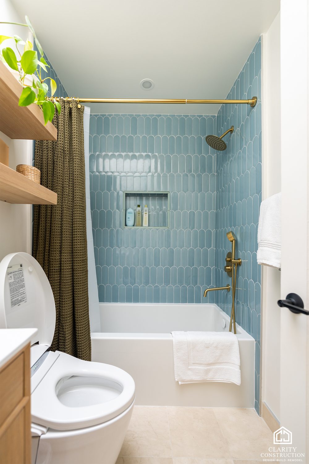 Compact bathroom with blue picket tile around the tub, brass fixtures and oak shelves