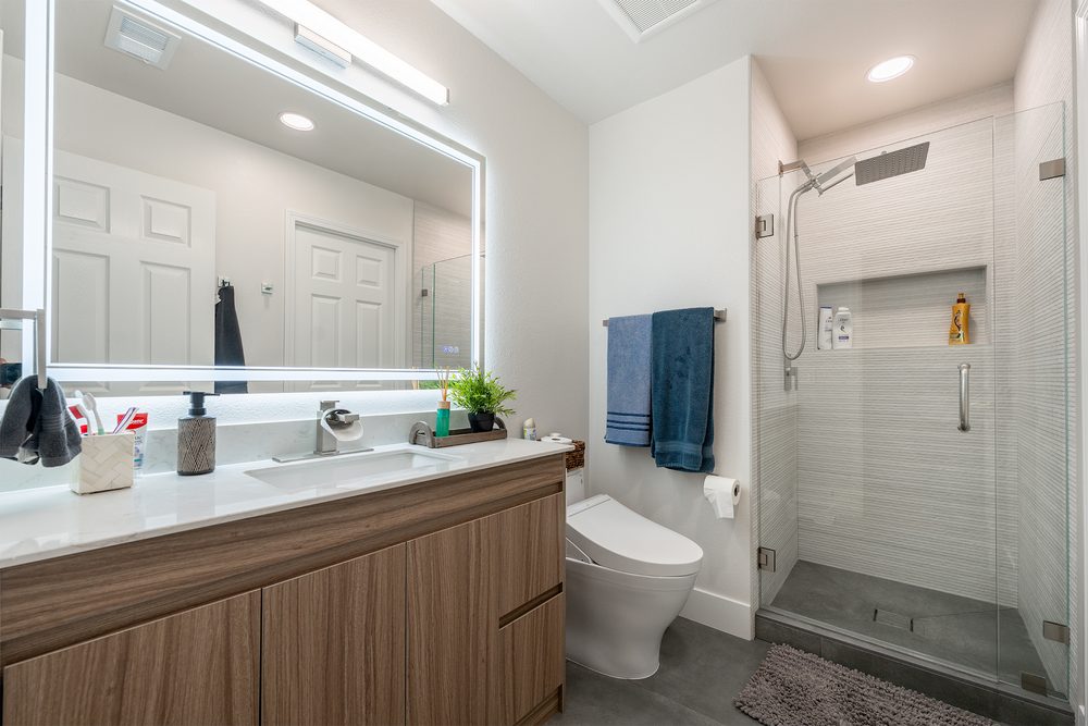 Hall bathroom with a walnut vanity, a backlit mirror and a white tile shower