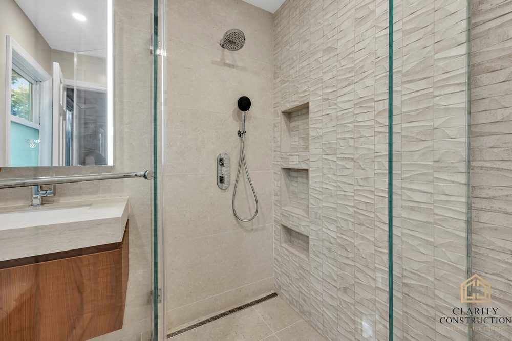Walnut vanity and backlit mirror beside the walk-in shower with its textured stone accent wall