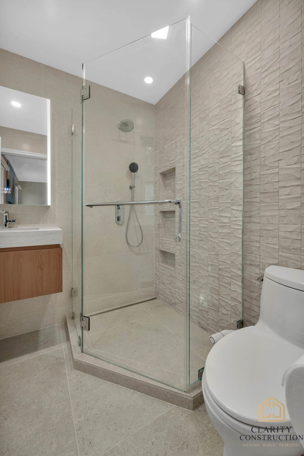 Frameless glass shower enclosure with a textured stone wall, beside the toilet and vanity